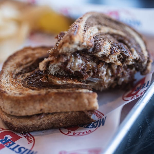 A grilled sandwich with melted cheese and caramelized onions, cut in half on Mister B's branded paper.