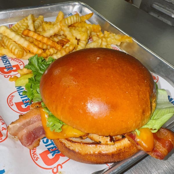 A chicken sandwich with bacon, cheese, and lettuce, served with crinkle-cut fries on a metal tray.