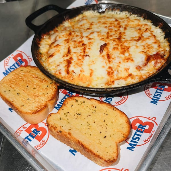 A cast iron skillet of cheesy dip with two slices of garlic bread on a Mister B's branded paper.