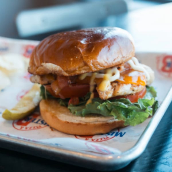 Mister B's grilled chicken sandwich with lettuce, tomato, and sauces, served with a pickle.