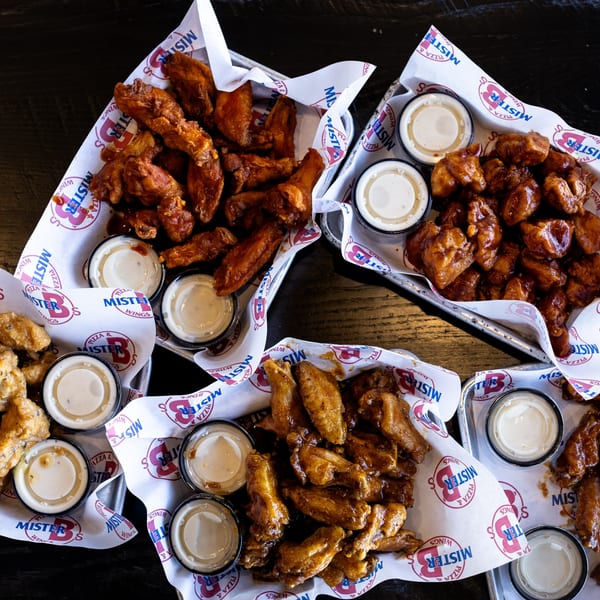 Four baskets of Mister B's wings in various flavors, with dipping sauces, on a dark table.