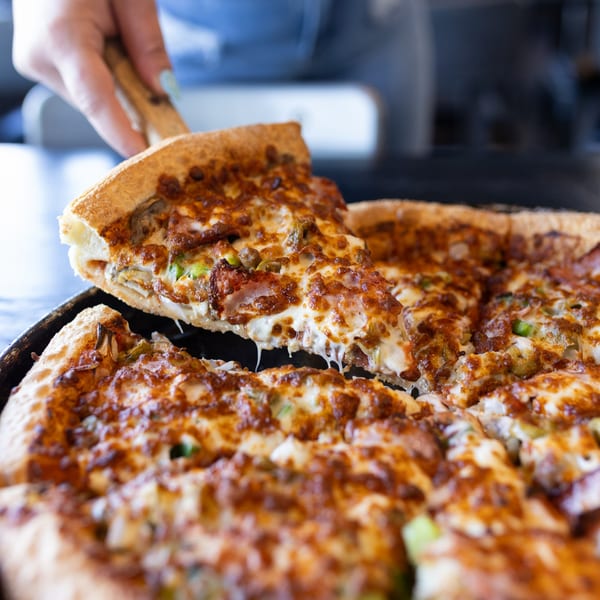 A hand lifts a slice of Mister B's Pizza, showing melted cheese and toppings.