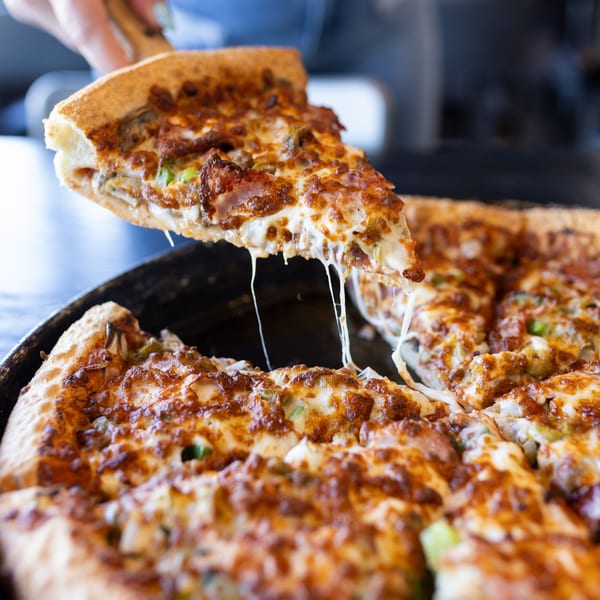 A hand lifts a slice of Mister B's Pizza with melted cheese stretching from the pan.