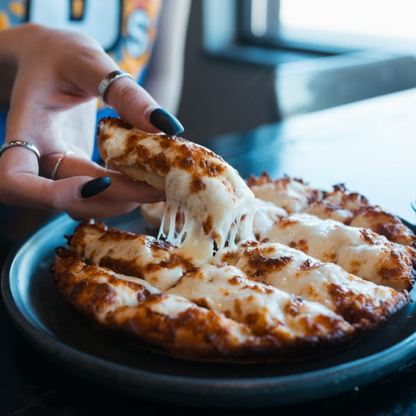 A hand with black nail polish pulls a cheesy breadstick from a Mister B's Pizza & Wings platter.