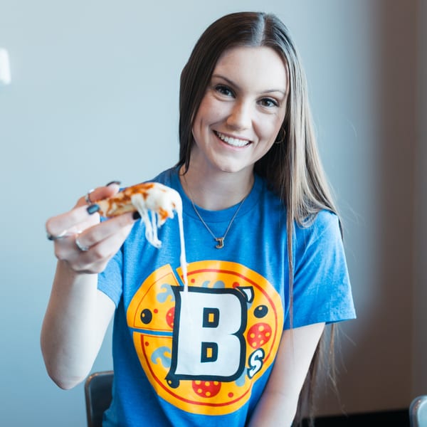 Smiling woman in a Mister B's Pizza & Wings t-shirt holding a cheesy slice of pizza.