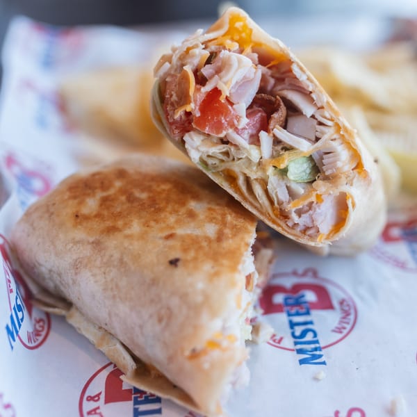Two halves of a grilled chicken wrap filled with chicken, tomatoes, and cheese on Mister B's branded paper.