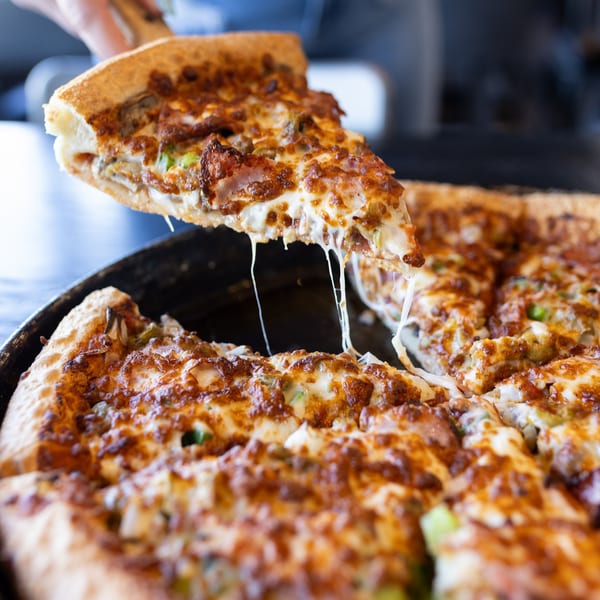A hand lifts a slice of Mister B's Pizza, with melted cheese stretching from the pan.